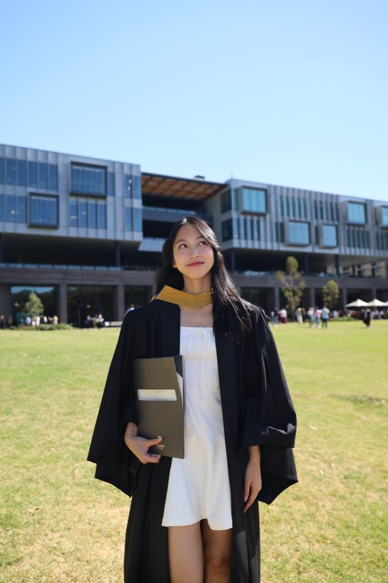 Graduate in gown and gold hood looking up in front of a modern campus building
