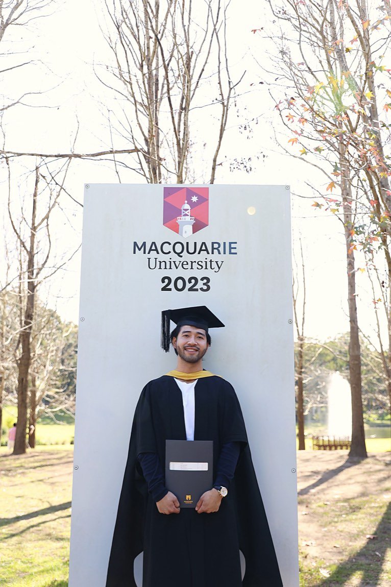 Graduate smiling with his testamur at the university sign